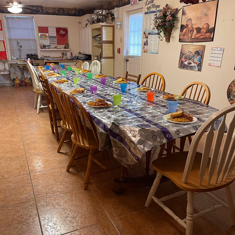 A long dining table covered with a silver and purple patterned tablecloth, set with plates of food and colorful plastic cups. The room has wooden chairs around the table, tiled floor, and walls decorated with framed pictures and a calendar. There is a door with a window and an exit sign above it in the background.
