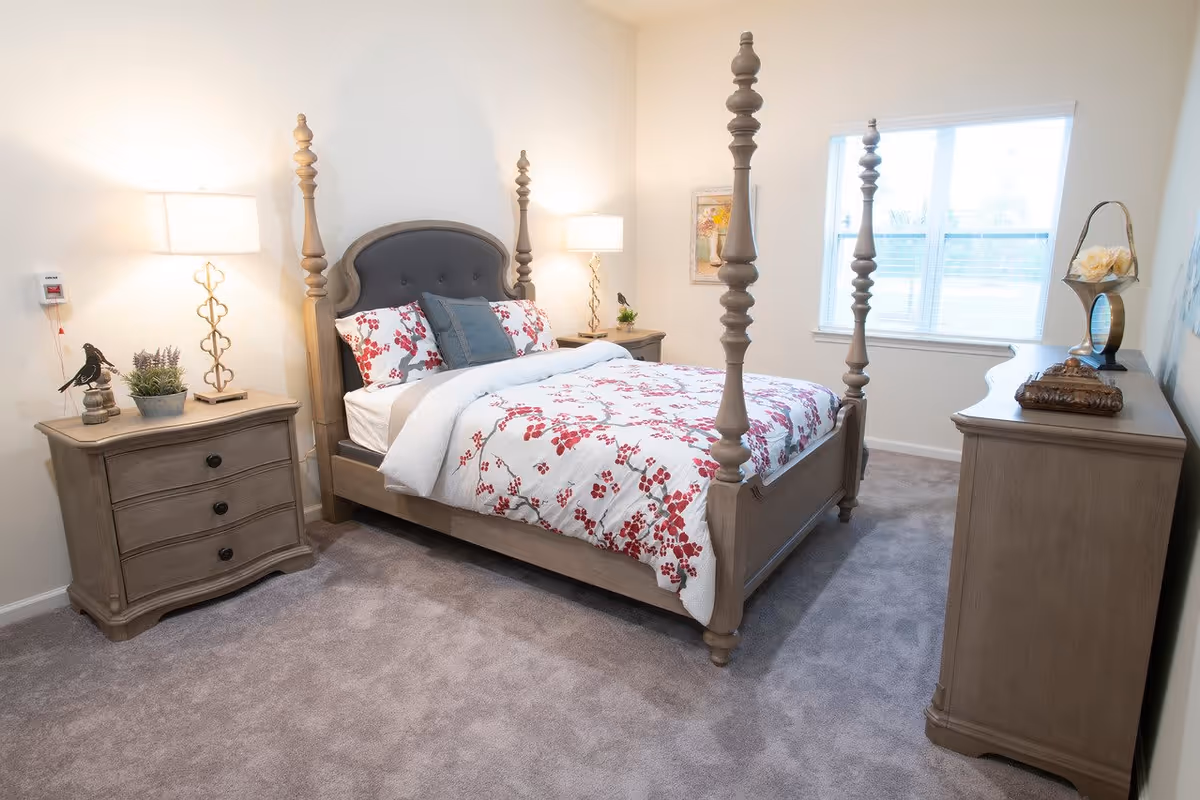 Well-lit bedroom with a four-poster bed dressed in floral bedding, matching wooden nightstands and dresser, and a window.