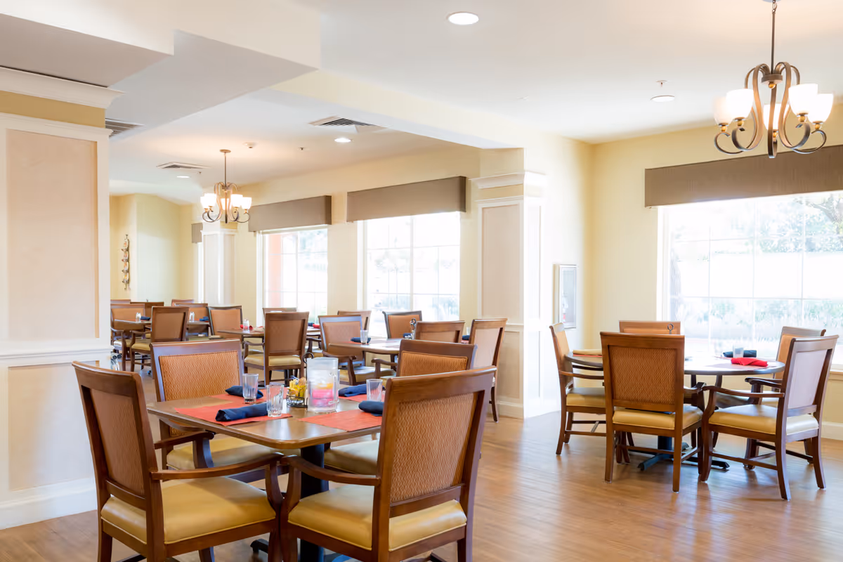 Bright dining room with wooden tables and chairs set for meals under chandeliers by large windows.