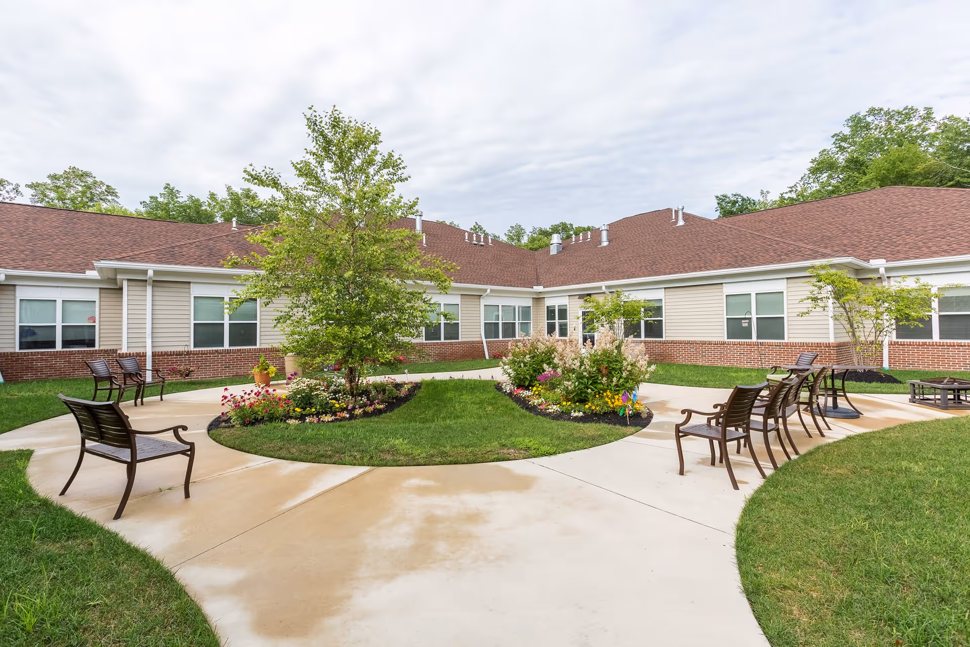 Outdoor courtyard area with a circular concrete walkway surrounding a grassy island with trees and flower beds, benches placed along the walkway, and a single-story building with beige siding and red brick exterior walls in the background under a cloudy sky.