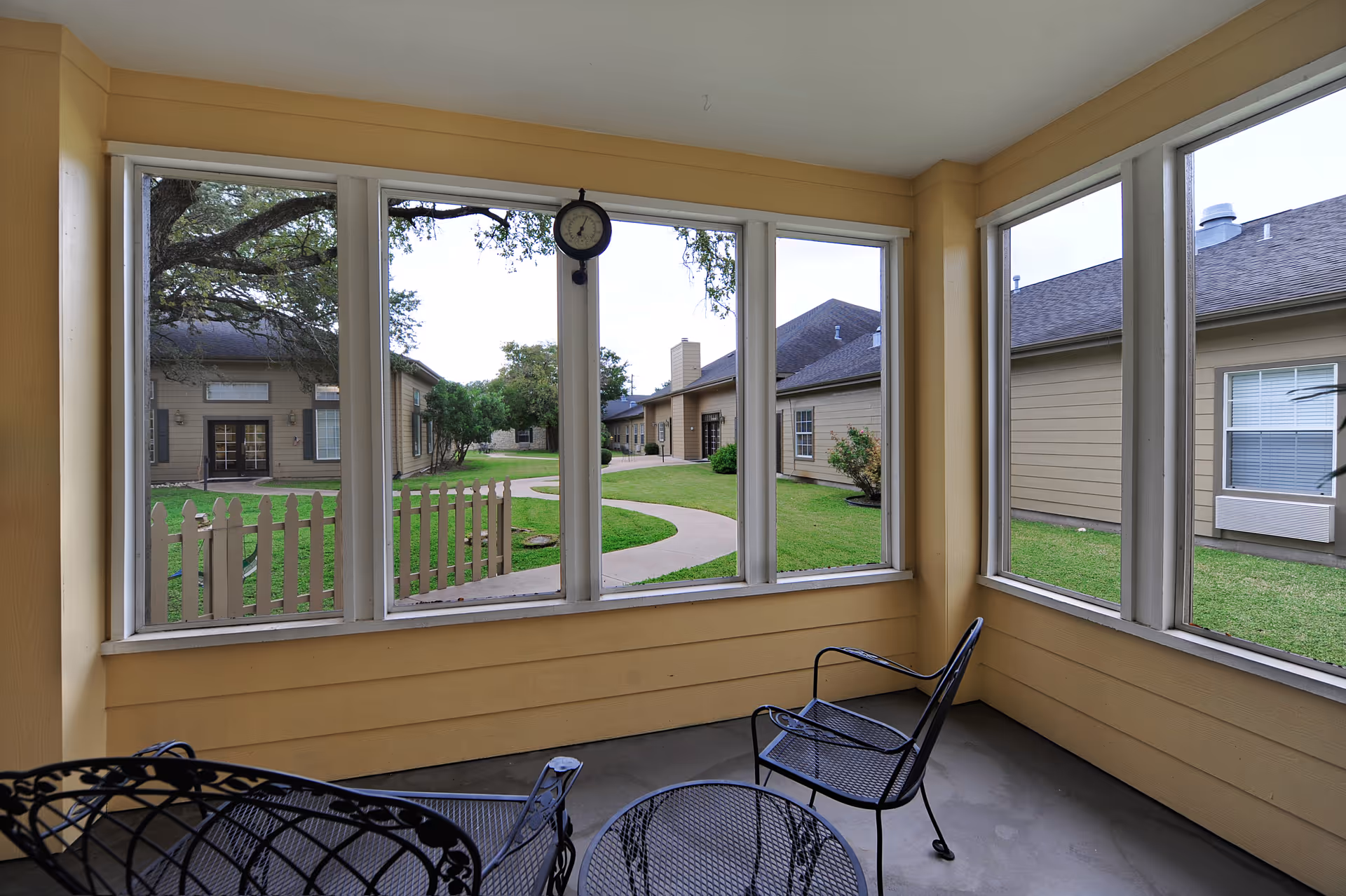 Enclosed porch with metal patio chairs and table looking out through large windows onto a grassy courtyard and surrounding single-story buildings.