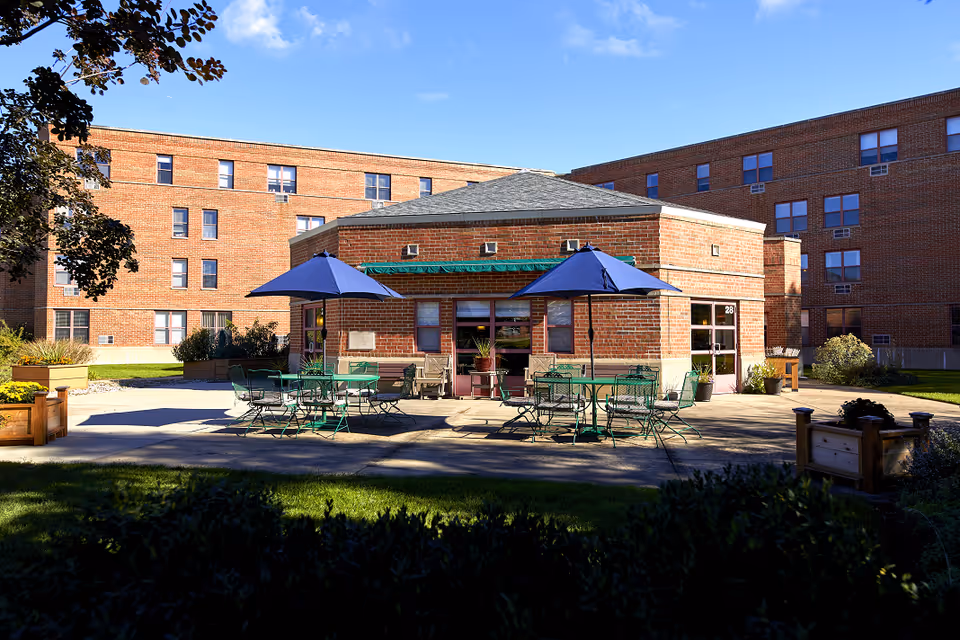 Outdoor patio area with green metal tables and chairs under blue umbrellas in front of a small brick building, surrounded by larger brick residential buildings under a blue sky with some clouds.