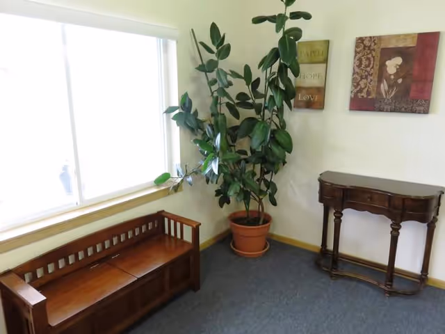 Corner of a sitting area featuring a wooden bench, potted plant, small console table, and wall art beside a large window.