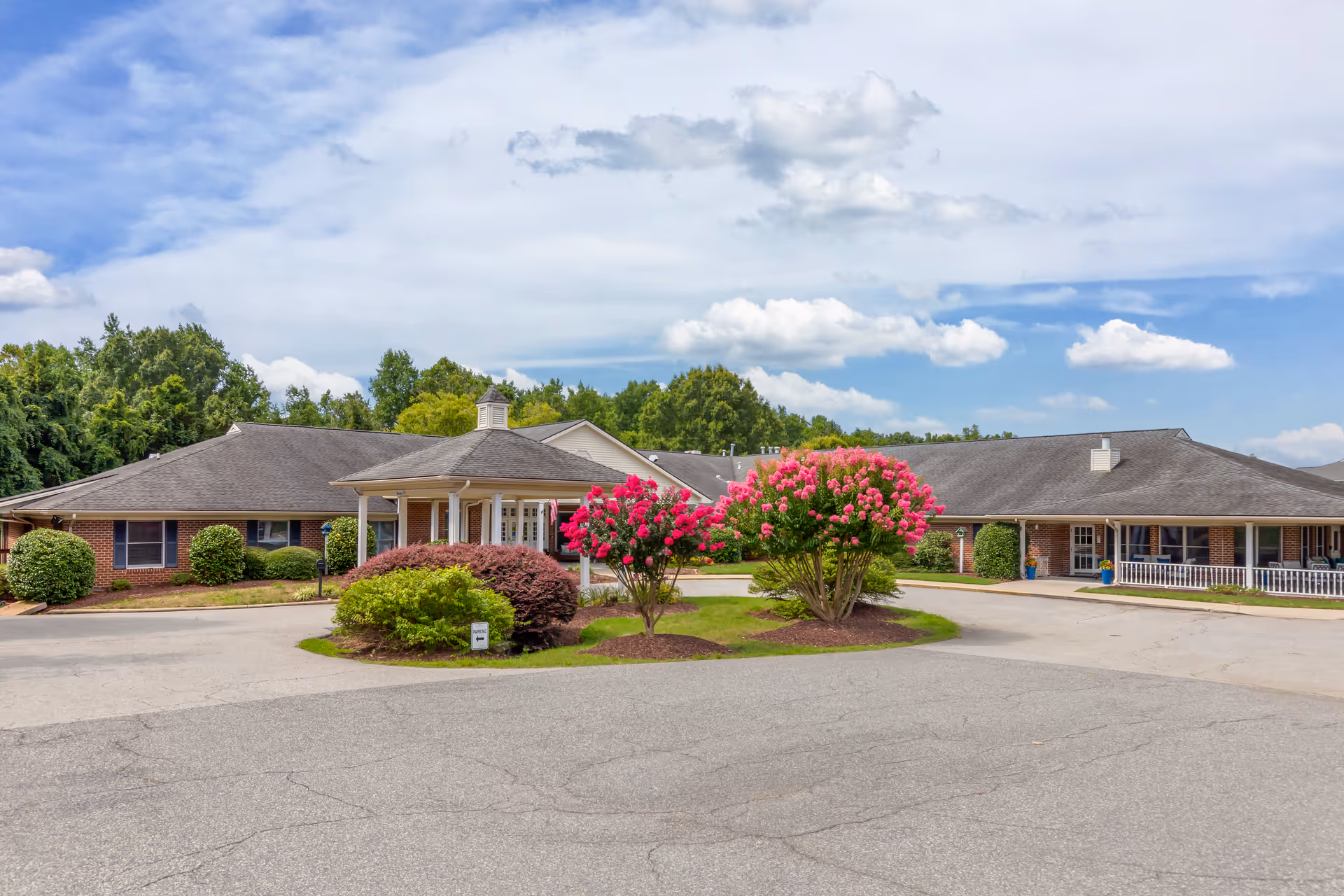 Exterior view of Brookdale High Point facility showing a single-story brick building with a covered entrance, surrounded by landscaped bushes and flowering trees under a partly cloudy sky.