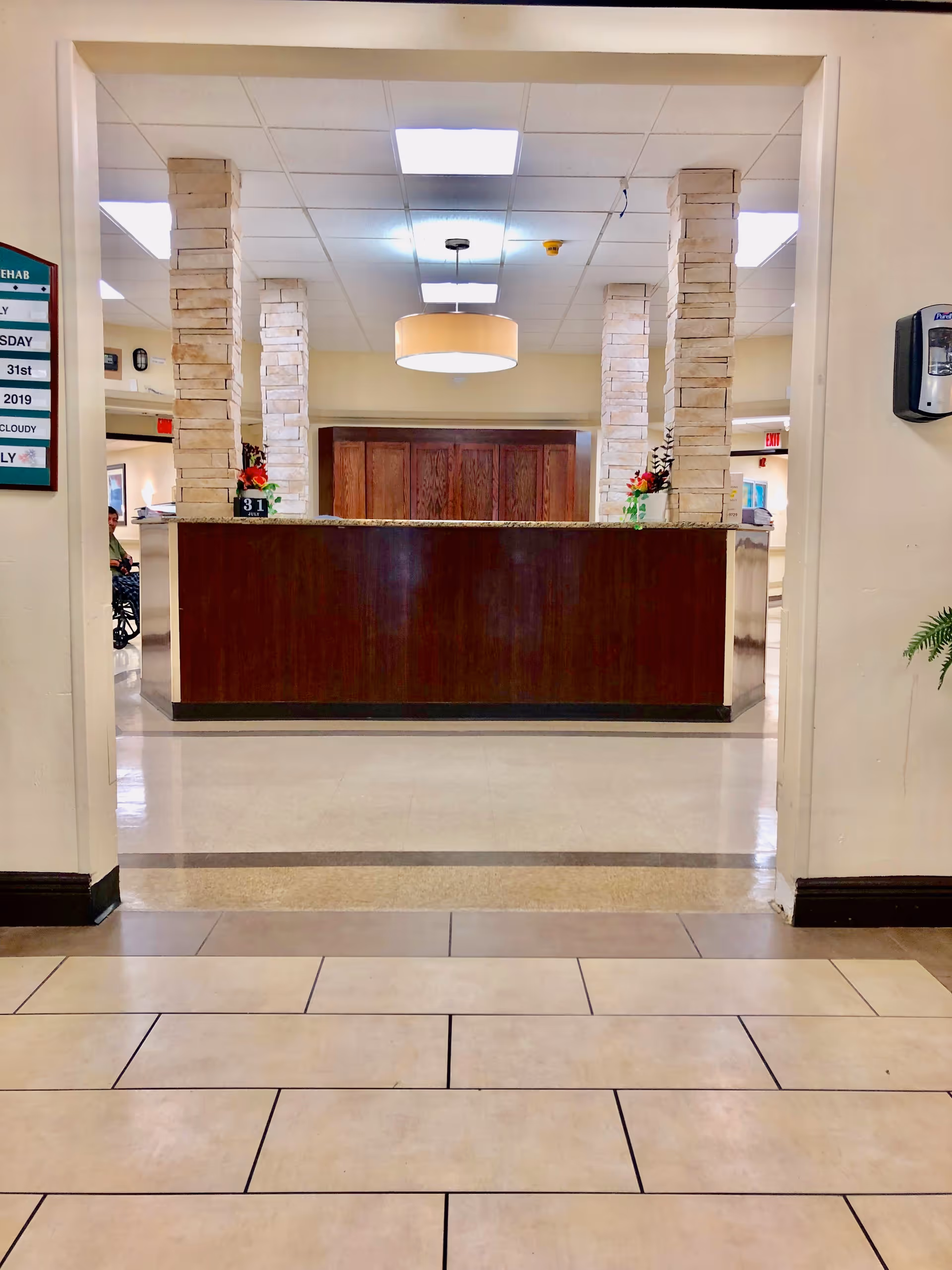 View of a reception desk area in a healthcare or rehabilitation facility with stone pillars, a hanging light fixture, and a tiled floor. There are flowers on the desk and a hand sanitizer dispenser mounted on the wall to the right.