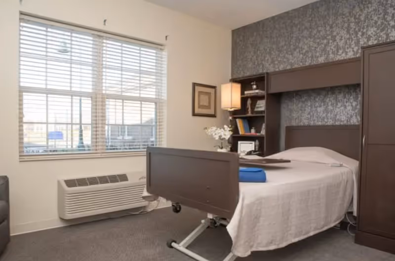 A single bed with a brown headboard and footboard in a room with a large window with blinds. Next to the bed is a built-in shelving unit with books, a lamp, and decorative items. The walls are light-colored with one wall featuring a patterned wallpaper. There is a heating or air conditioning unit below the window.