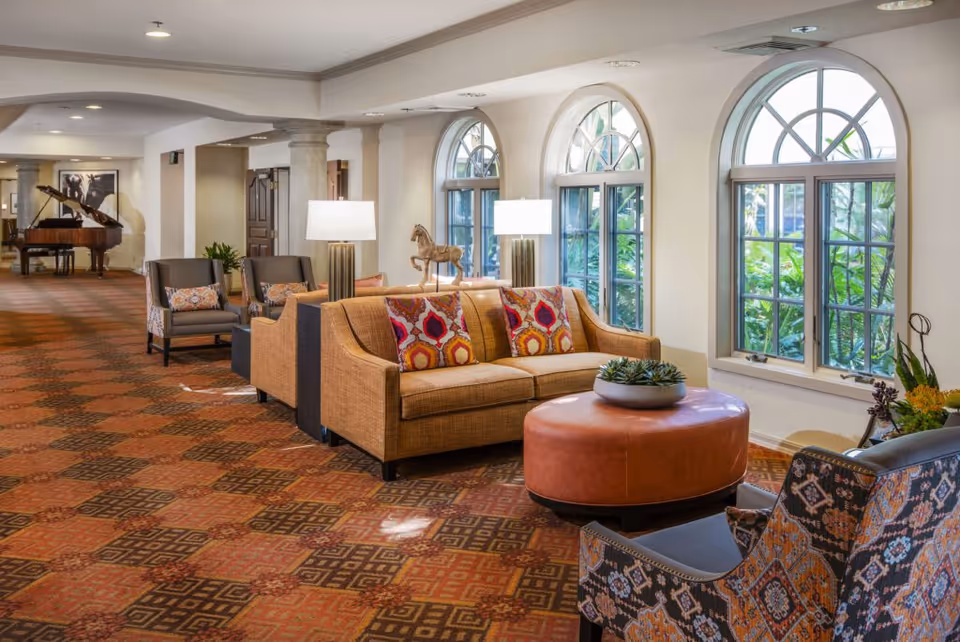 A spacious and well-lit lounge area with a patterned carpet, a tan sofa with colorful cushions, two patterned armchairs, a round leather ottoman with a plant on top, and large arched windows letting in natural light. In the background, there is a grand piano and additional seating along the hallway.