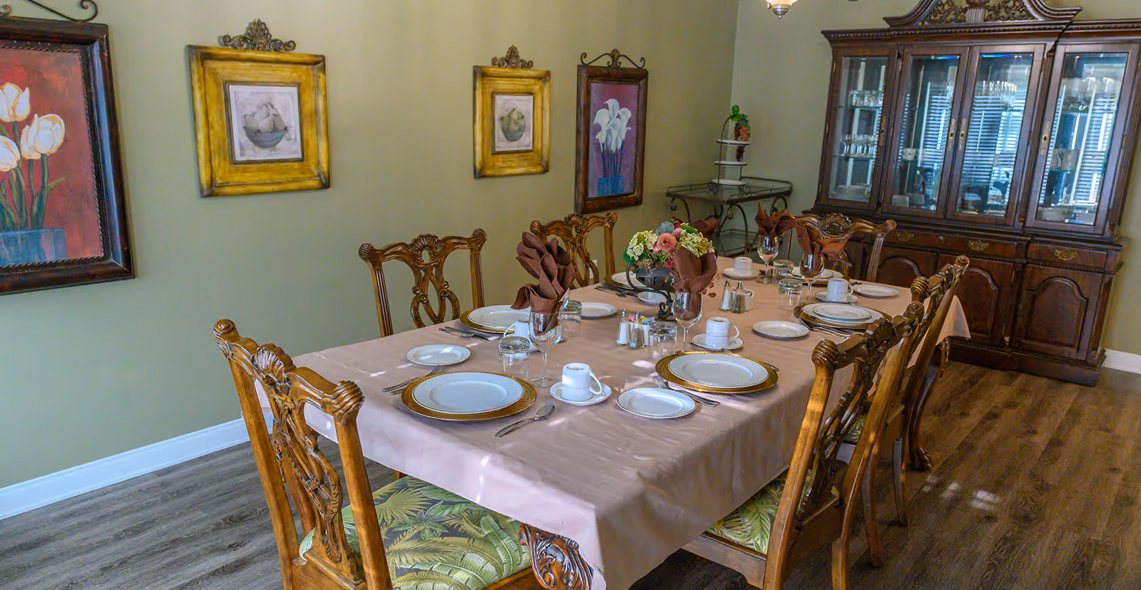 A formal dining room with a long rectangular table covered with a beige tablecloth. The table is set with white plates, gold-rimmed chargers, cups, glasses, silverware, and brown folded napkins. A floral centerpiece is in the middle of the table. The room has wooden chairs with ornate backs and green patterned cushions. On the walls are framed paintings of flowers and fruit. A large wooden china cabinet filled with glassware is against the far wall, and a small metal serving cart is in the corner.