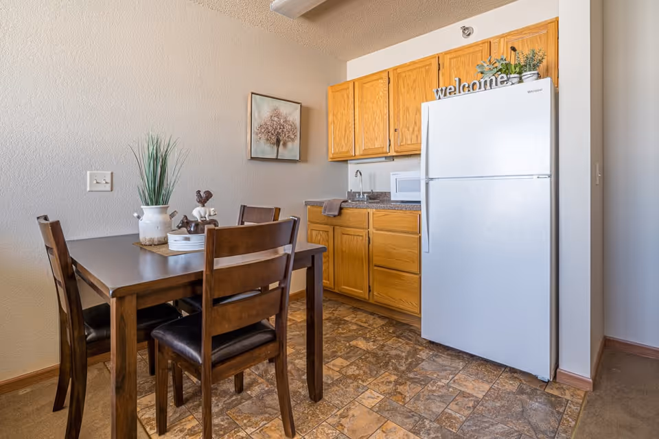 A small kitchen and dining area with a wooden table and four chairs. The kitchen has wooden cabinets, a white refrigerator, a microwave, and a sink. There is a decorative plant and a 'welcome' sign on top of the refrigerator, and a framed picture of a tree on the wall.