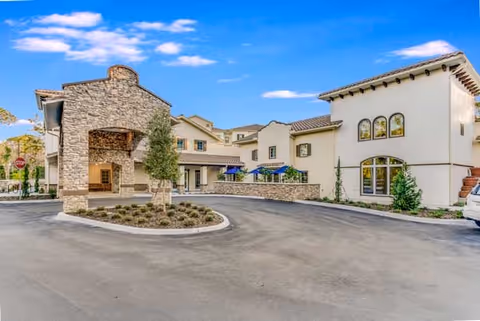 Exterior view of Tuscan Gardens of Palm Coast Senior Living facility showing a large stone entrance with a covered drop-off area, surrounded by a circular driveway and landscaped with small bushes and trees under a blue sky with some clouds.