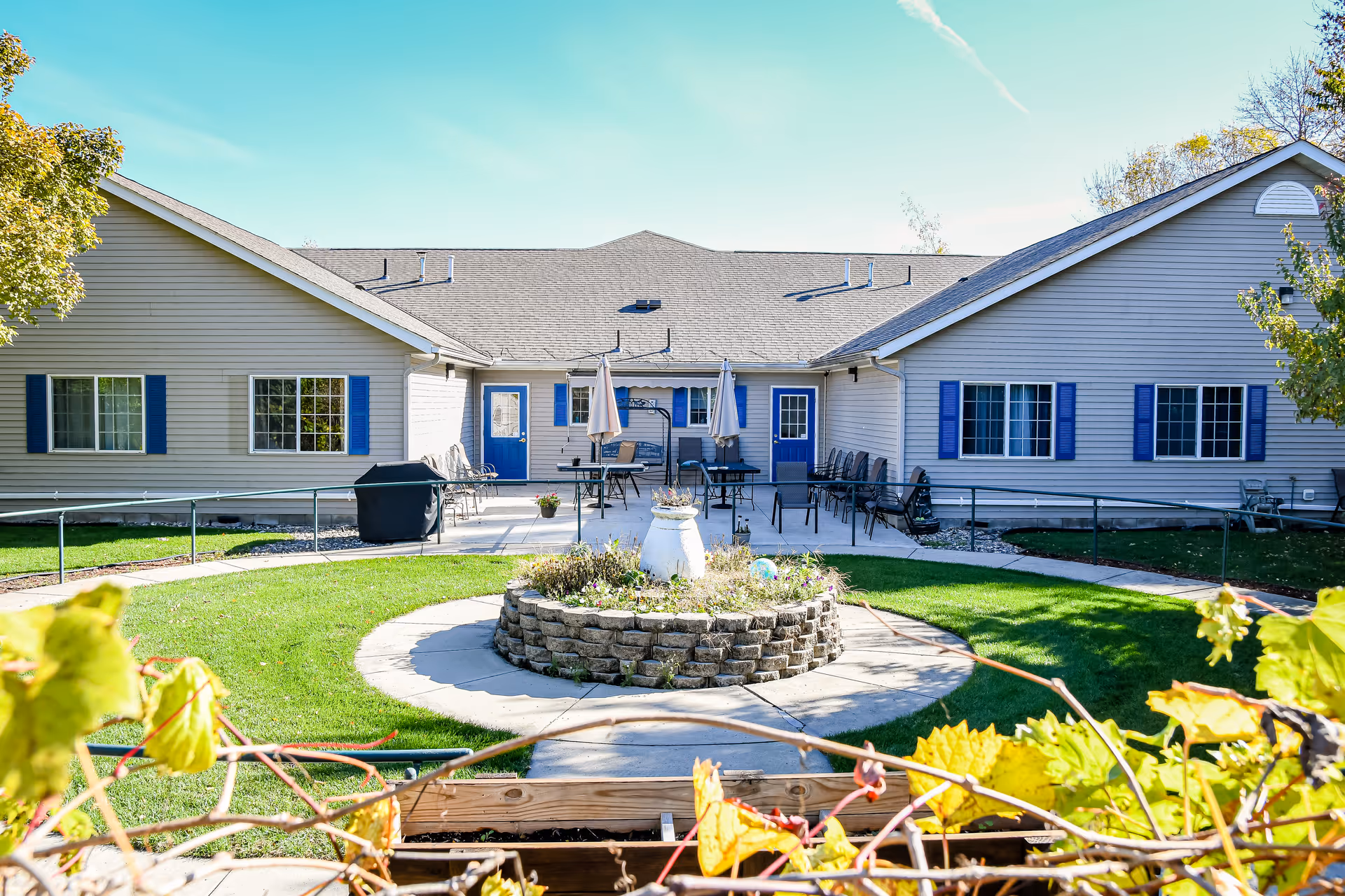 Front courtyard of a single-story senior living building with a circular raised flowerbed, patio seating, and blue-trim windows and doors.