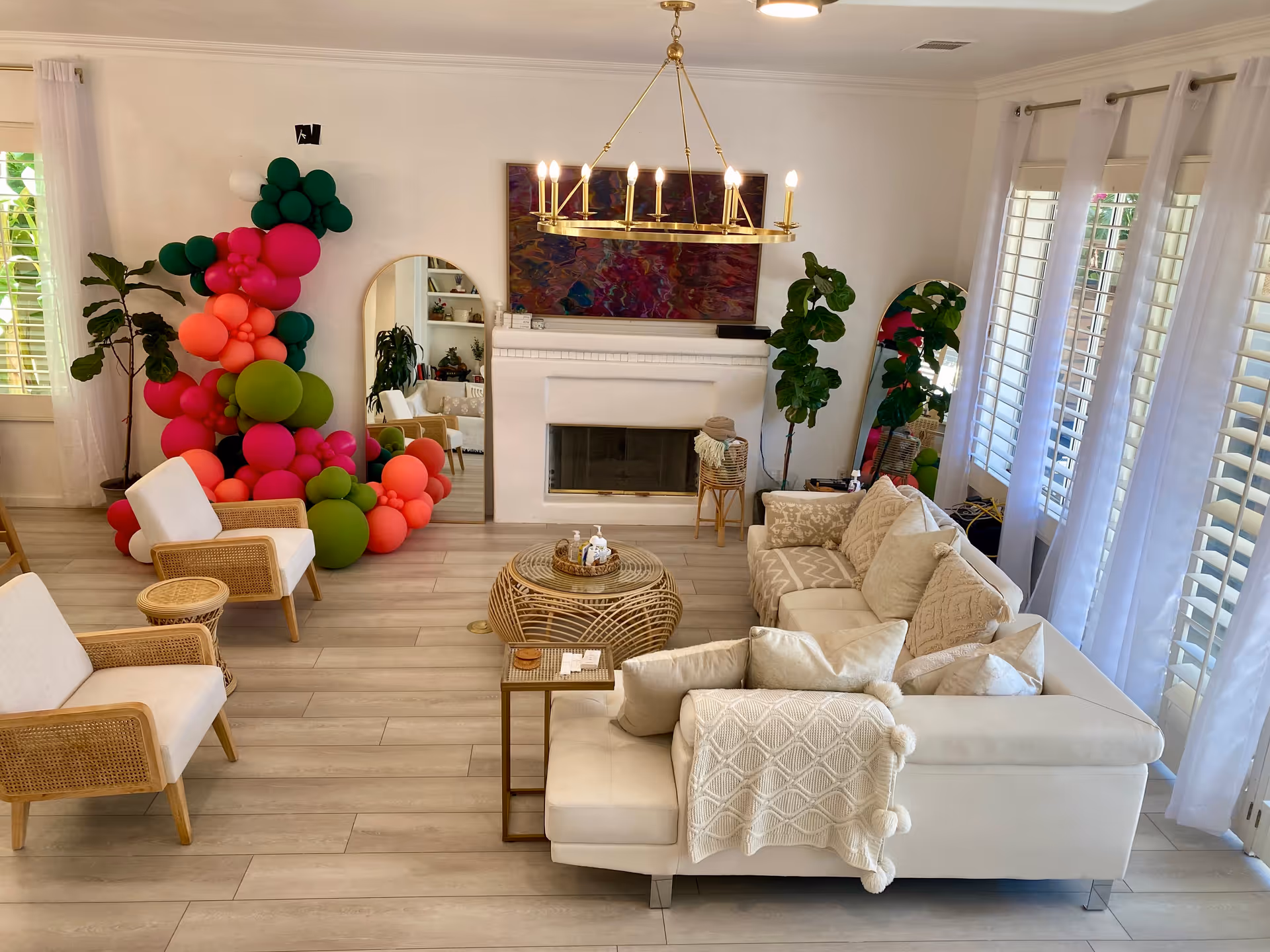 Bright living room with a white sectional and armchairs facing a fireplace, a colorful balloon display by a mirror, and large shuttered windows.