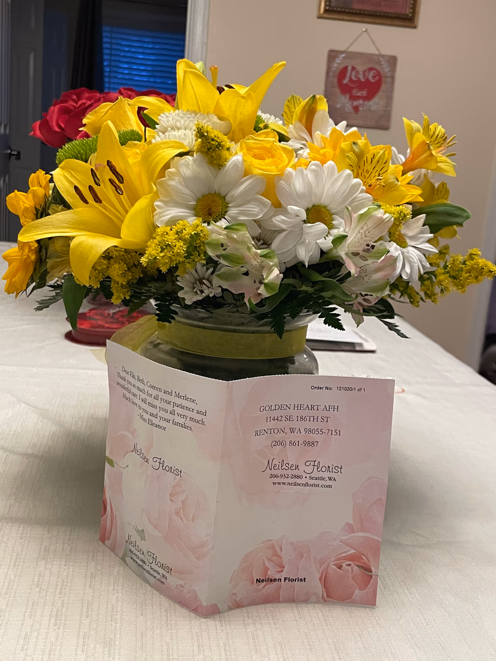 A bouquet of yellow lilies, white daisies, and other yellow and white flowers arranged in a glass vase with a green ribbon, placed on a white tablecloth. In front of the vase is a card from Neilsen Florist addressed to Golden Heart Adult Family Home in Renton, WA. The background shows a beige wall with a framed picture and a hanging decoration with the word 'Love'.