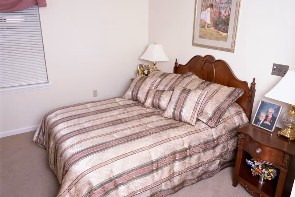 A bedroom with a wooden bed frame and a bed made with a striped comforter and matching pillows. There are two bedside tables, each with a lamp and decorative items including a framed photo and a small flower arrangement. A window with closed blinds and a valance is on the left wall, and a framed picture hangs above the bed.