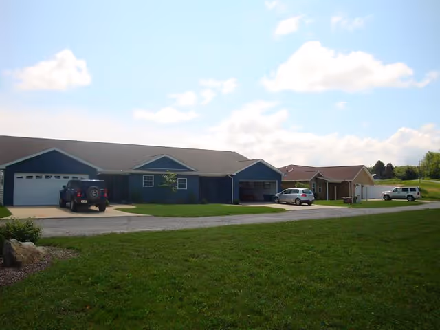 Single-story residential buildings with attached garages, parked cars, and a large grassy area in the foreground under a partly cloudy sky.