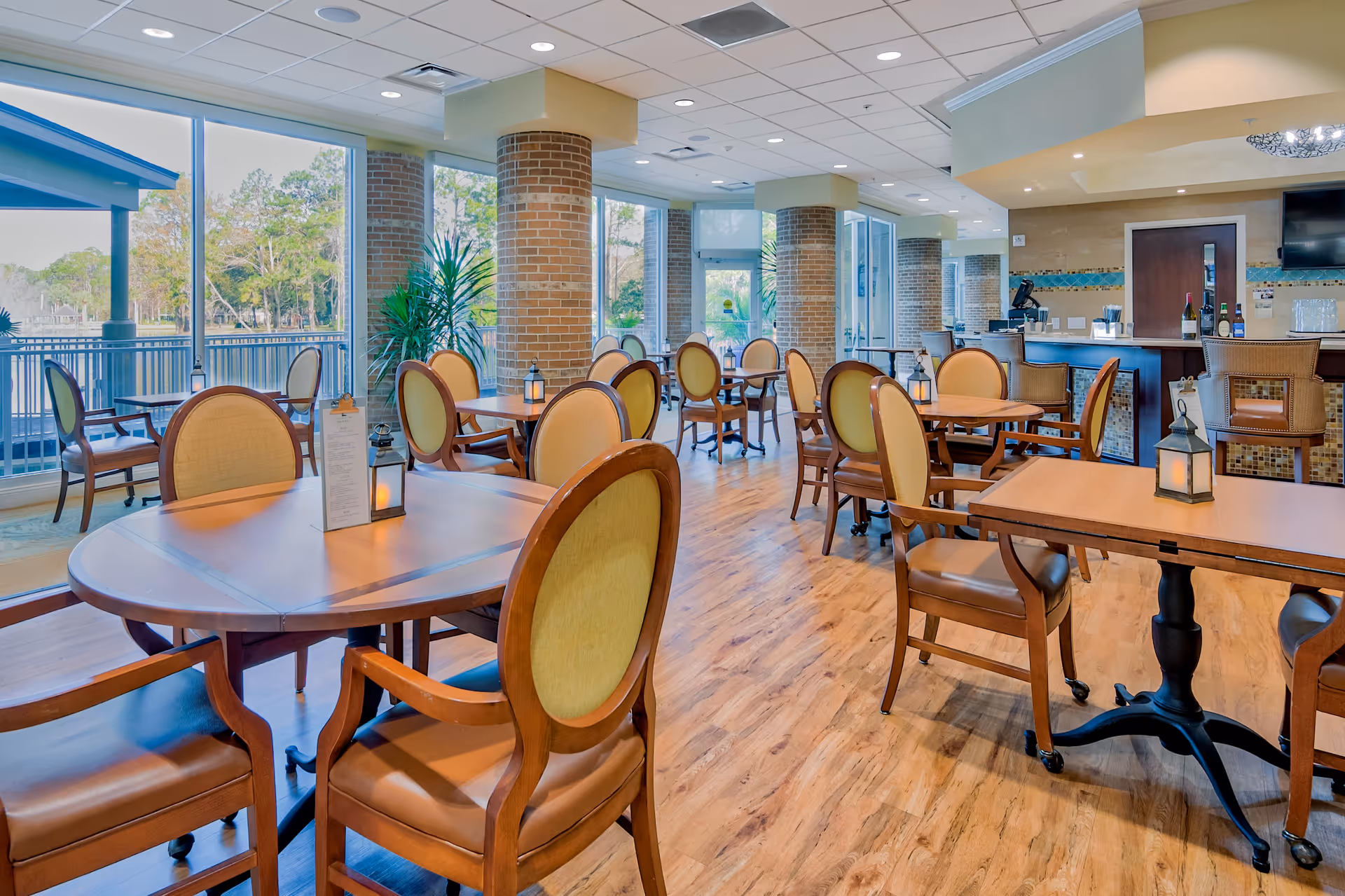 Spacious, sunlit dining room with round and rectangular wooden tables and upholstered chairs next to large windows and a serving counter.