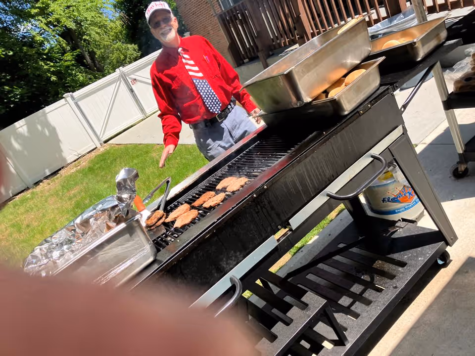 An elderly man wearing a red shirt, patriotic tie, and a white cap is standing outdoors next to a large grill with several hamburger patties cooking. There are trays with buns and other items on the grill. The setting is a sunny day with a white fence and green grass in the background.