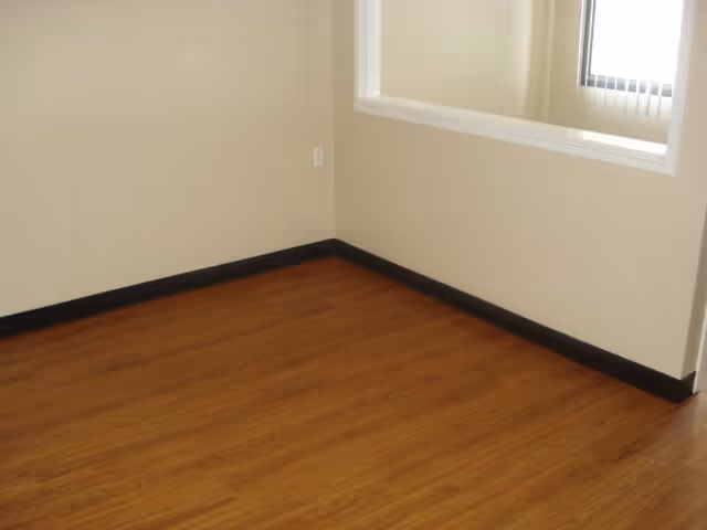 Empty room with light beige walls, a window with vertical blinds, and wood-look flooring with dark baseboards.