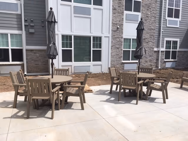 Outdoor patio area with two round tables, each surrounded by four wooden chairs. Each table has a closed black umbrella. The patio is adjacent to a building with gray and white siding and multiple windows.