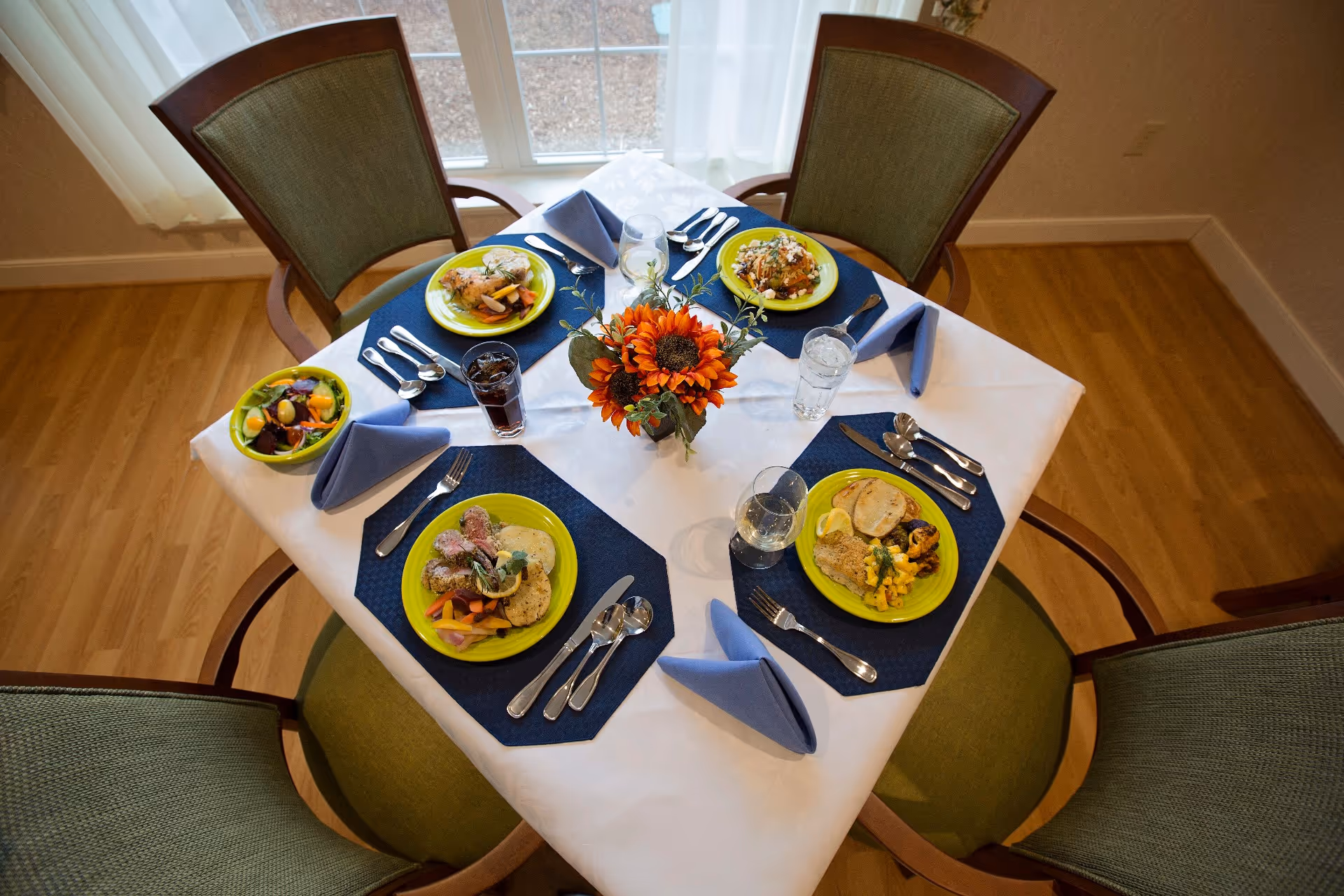A dining table set for four with green cushioned chairs around it. The table is covered with a white tablecloth and blue placemats, each with a neatly folded blue napkin and silverware. Each place setting has a plate with a different meal, and there are glasses of water and soda. A centerpiece with orange sunflowers is in the middle of the table. The background shows a window with sheer white curtains.