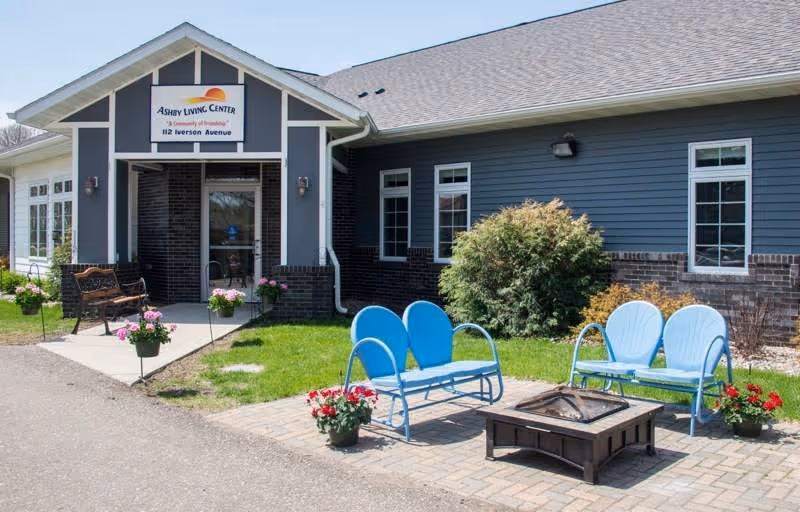 Exterior view of Ashby Living Center showing the entrance with a sign above the door. The building has blue siding with brick accents. In front of the entrance, there is a paved patio area with blue metal outdoor seating and a fire pit surrounded by flower pots. The area is landscaped with grass, bushes, and flowers.