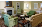 A cozy living room with a beige sofa, two green armchairs, a wooden coffee table, and a fireplace with a painting above it. The room has light yellow walls, a patterned carpet, and decorative items on the tables and shelves.