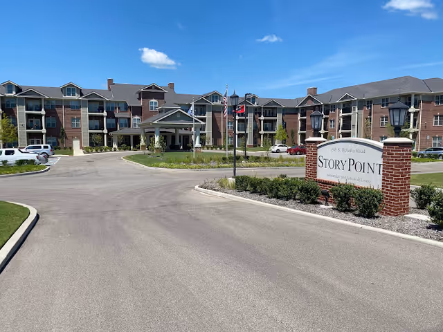 Exterior view of StoryPoint Collierville, a large multi-story senior living facility with a circular driveway, landscaped greenery, and a sign displaying the facility's name and address under a clear blue sky.
