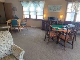 A cozy senior living common area with carpeted floor, featuring a patterned armchair in the foreground, a wooden table with four chairs, and another table with a green felt surface and four chairs. The room has large windows with sheer curtains allowing natural light, a sideboard with lamps and a stereo system, and framed artwork on the wall.