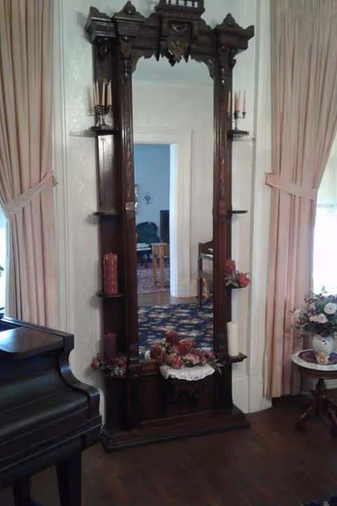 A tall, ornate wooden mirror with shelves holding candles and floral decorations, positioned between two windows with pink curtains in a room with wooden flooring. A black piano is partially visible on the left, and a small round table with a floral arrangement is on the right.