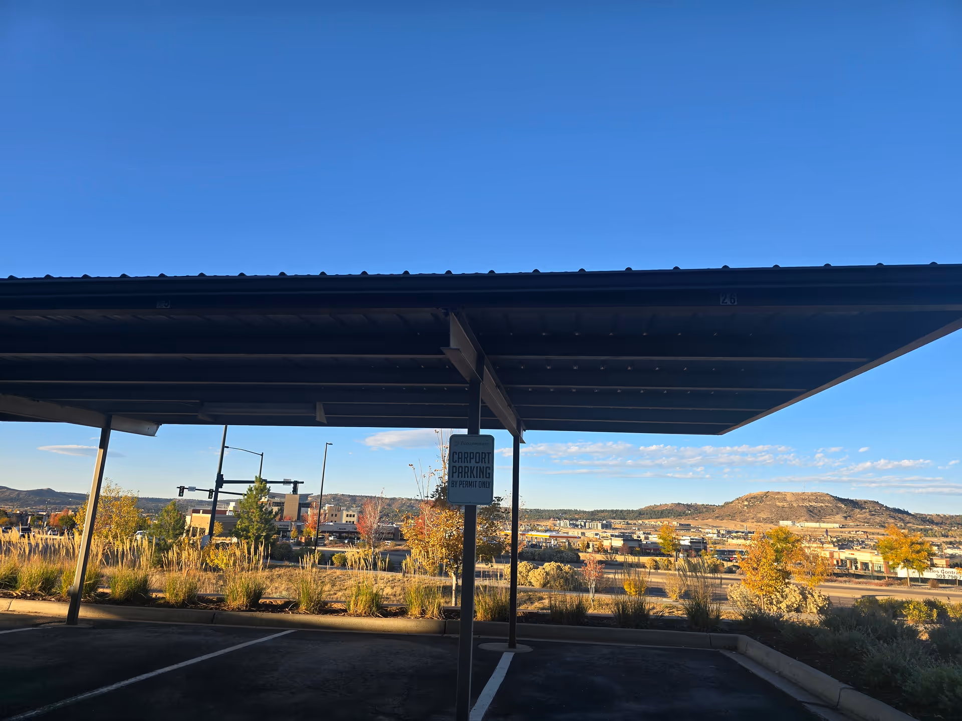 Covered carport parking area with a sign that reads 'Bonaventure Carport Parking By Permit Only,' overlooking a landscape with trees, buildings, and a hill under a clear blue sky.