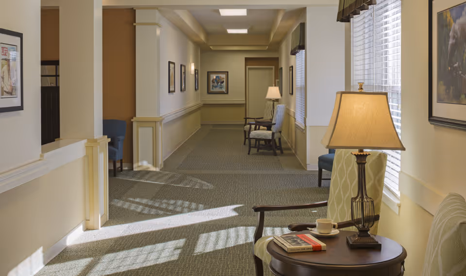 A well-lit hallway in a senior living facility with beige walls and carpeted floor. Several chairs are placed along the walls, with framed artwork hanging above them. A small round table with a lamp, a cup and saucer, and two books is positioned near a window with blinds partially open, casting shadows on the floor.