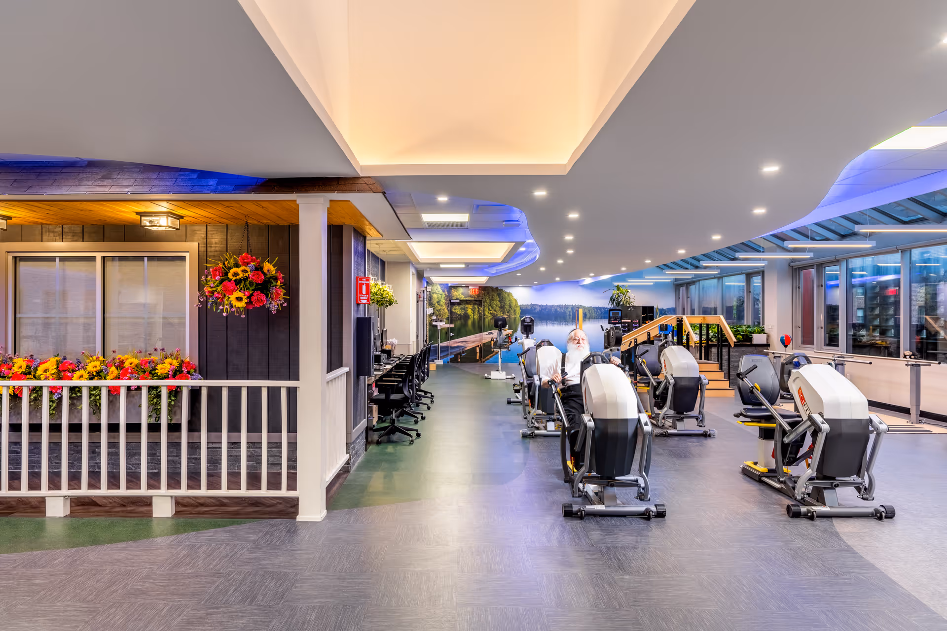 A spacious rehabilitation and exercise room with several exercise machines arranged in rows. A man with a white beard is using one of the machines. The room has large windows on the right side, allowing natural light to enter. On the left side, there is a decorative area with flower boxes and hanging flower baskets, resembling a porch. The ceiling has modern recessed lighting and a blue accent light. The far wall features a large mural of a serene lake scene with trees and a dock.