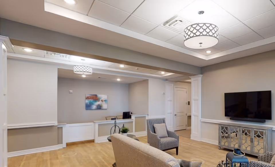 A modern senior living facility common area with beige walls and light wood flooring. The room features a flat-screen TV mounted on the wall above a decorative cabinet, a patterned armchair, a beige sofa, and a small round table with a plant and decorative items. The ceiling has recessed lighting and a stylish pendant light fixture. There is a reception desk area in the background with a piece of abstract artwork on the wall.