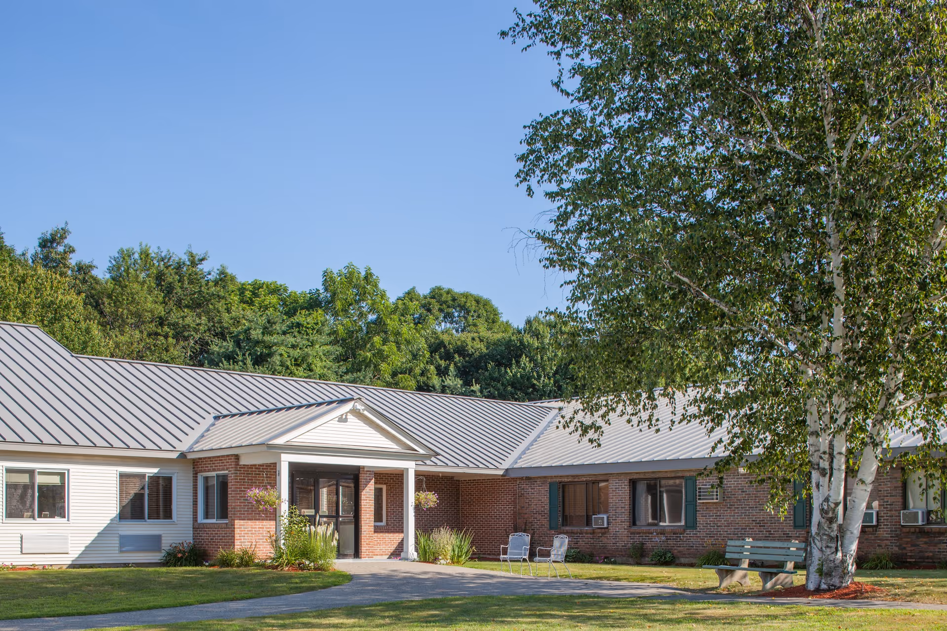 Exterior view of a single-story brick and siding building with a metal roof, surrounded by green trees and a well-maintained lawn. There is a paved walkway leading to the entrance, which has a small covered porch with white columns. Outdoor chairs and a bench are placed near the building under a large tree.