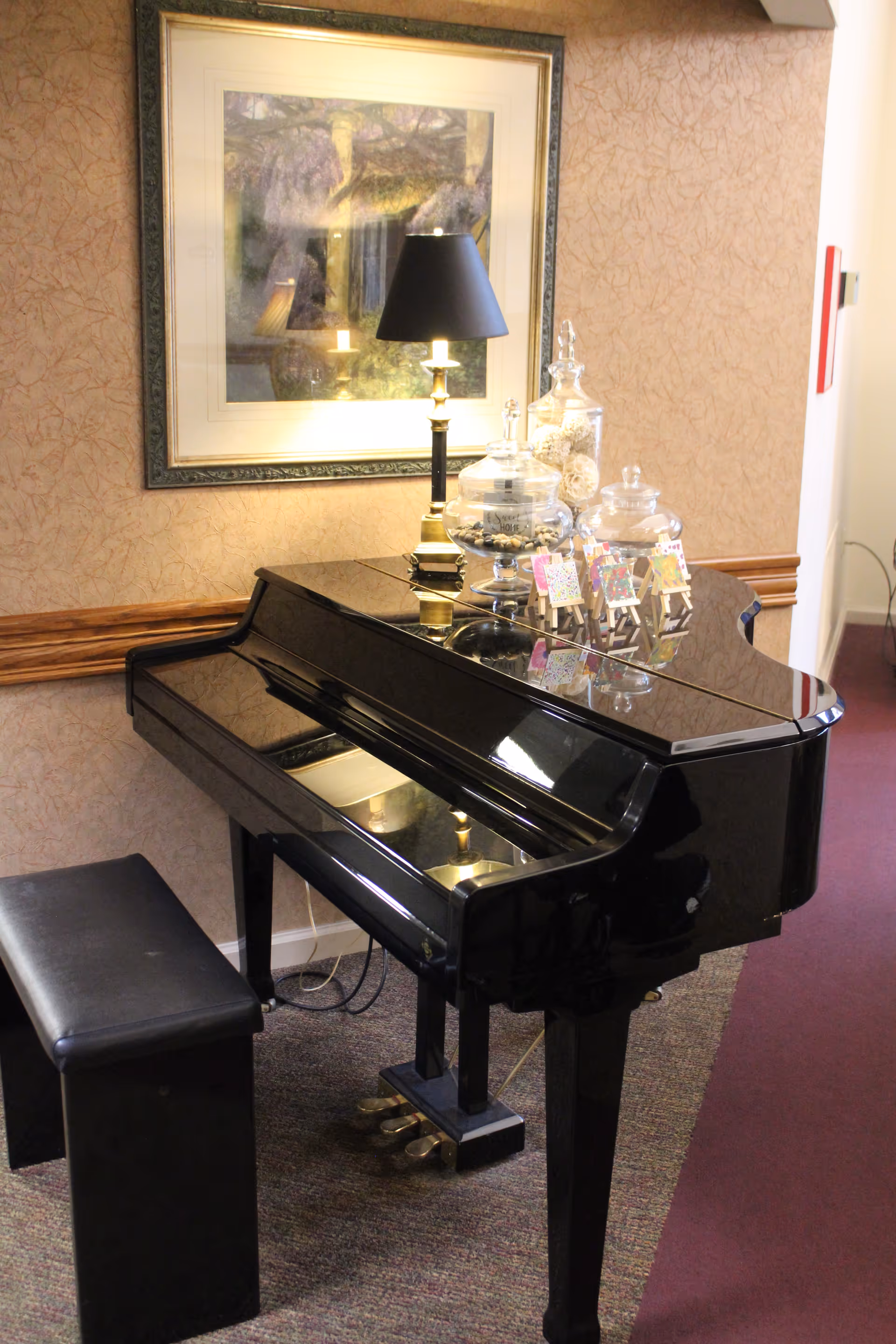 A black grand piano with a matching bench in a carpeted room. On top of the piano are decorative glass jars and a small black table lamp. A framed artwork hangs on the wall behind the piano.