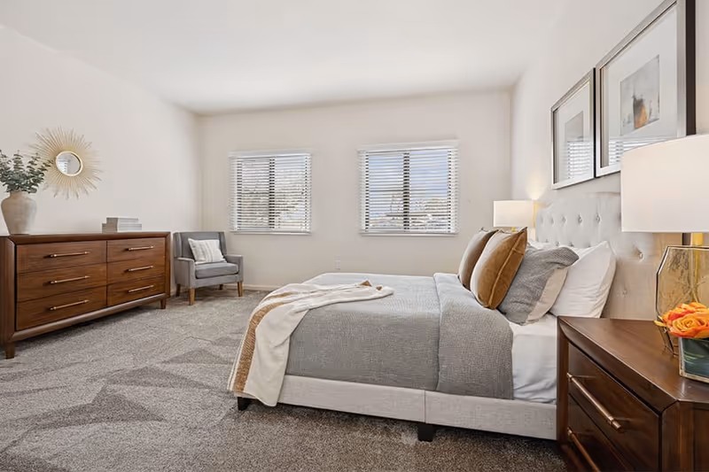 A bright and spacious bedroom with a large bed featuring gray bedding and multiple pillows. There are two windows with blinds letting in natural light. The room includes a wooden dresser with decorative items and a gray armchair in the corner. Two framed artworks hang above the bed, and a bedside table with a lamp and a small vase of orange flowers is visible.