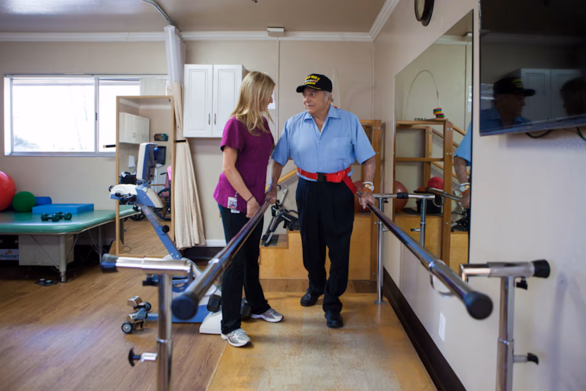 An elderly man wearing a blue shirt and black pants is using parallel bars for walking exercise in a rehabilitation or physical therapy room. A female caregiver or therapist in a purple shirt is standing next to him, providing support and encouragement. The room has wooden floors, exercise equipment, a large mirror on the wall, and a window letting in natural light.