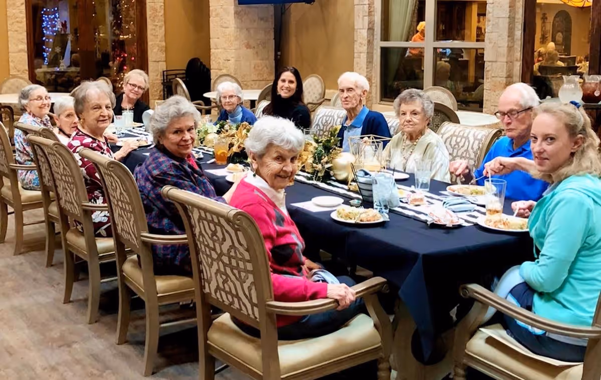 A group of elderly people and two younger adults sitting around a long dining table in a well-lit room, enjoying a meal together. The table is decorated with a black tablecloth and a centerpiece with gold ornaments. The setting appears warm and communal.