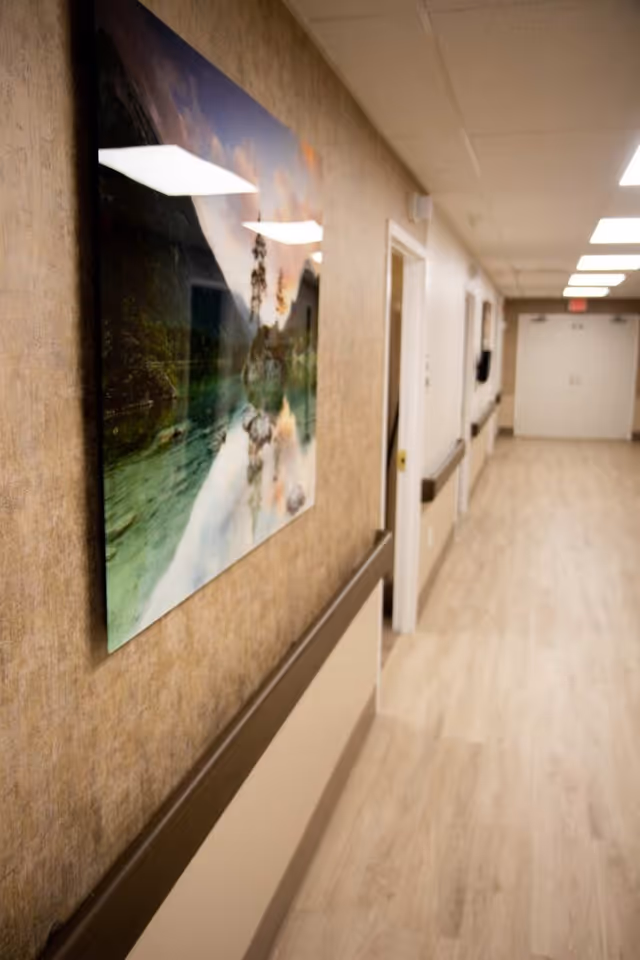 Well-lit interior hallway with wood-look flooring, handrails, wall art, and doors leading to rooms.