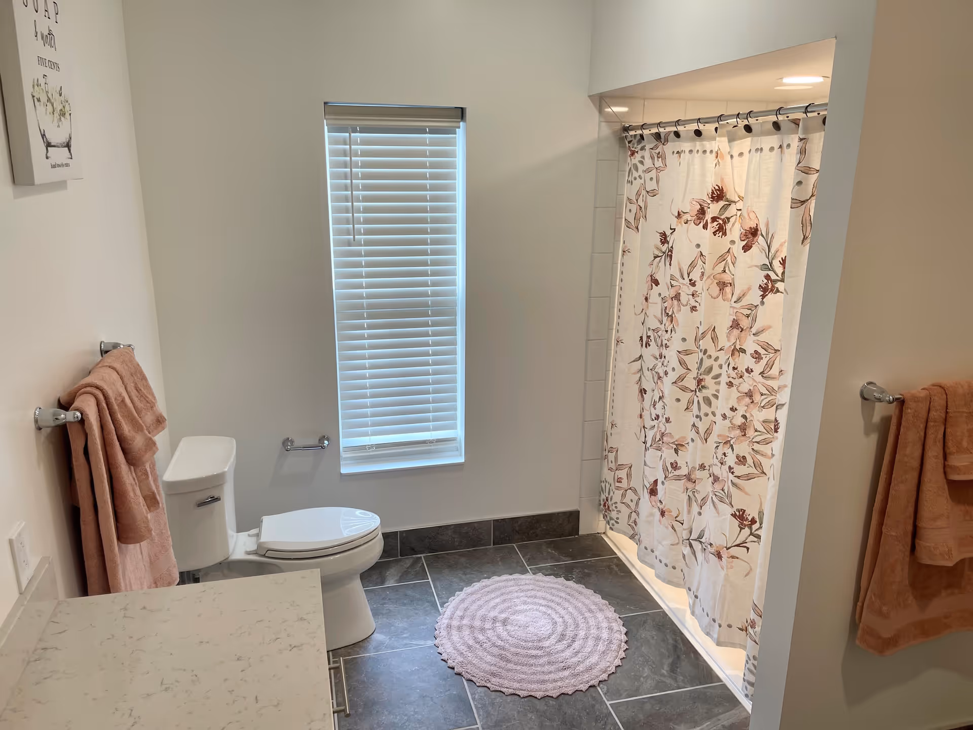 A clean bathroom with a white toilet, a window with closed blinds, a shower with a floral-patterned curtain, pink towels hanging on towel racks, a round pink bath mat on the dark tiled floor, and a white countertop partially visible.