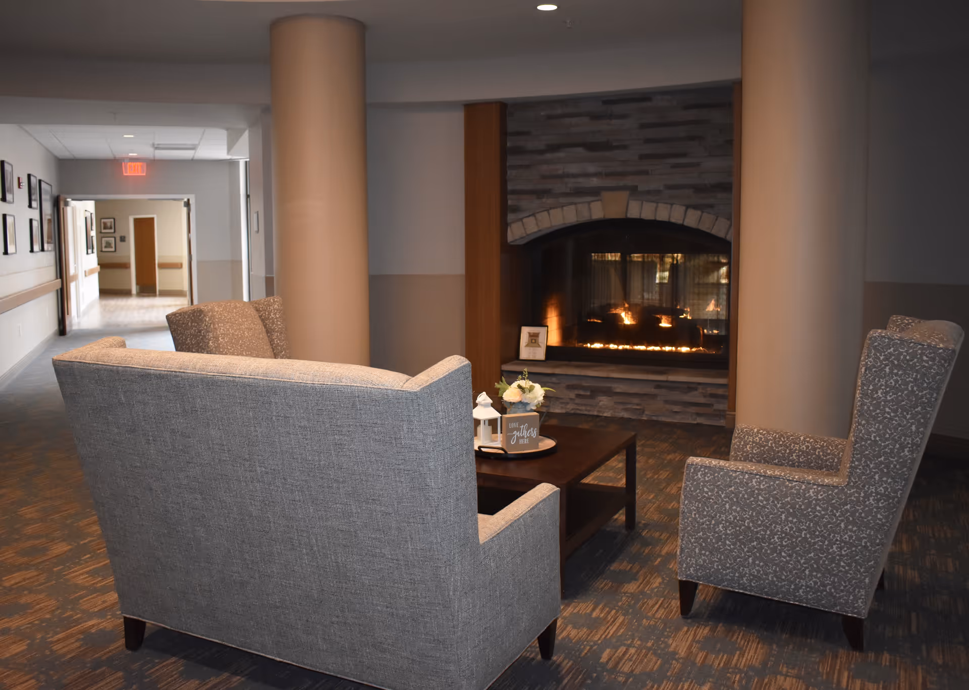 A cozy seating area in a senior living facility with a gray couch and two patterned armchairs arranged around a wooden coffee table. The table holds a small decorative lantern, a flower arrangement, and a sign that says 'Love gathers here.' In the background, there is a stone fireplace with a warm fire burning. The area is flanked by two large beige columns and a hallway is visible to the left.