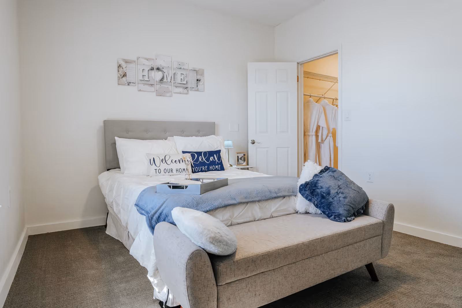 A neatly made bed with white linens and decorative pillows in a bedroom. Above the bed is wall art spelling 'HOME'. A small bench with cushions is placed at the foot of the bed. An open door reveals a closet with hanging robes inside. The room has light-colored walls and carpeted flooring.