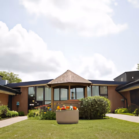 Exterior view of a single-story building with large windows and a small covered porch area. The building is surrounded by green grass, bushes, and a paved walkway. The sky is partly cloudy.