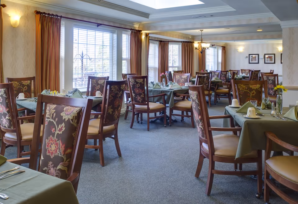 A spacious dining room with multiple tables covered in green tablecloths, each set with cups, napkins, and silverware. The chairs have floral patterned upholstery and wooden frames. Large windows with striped curtains allow natural light to fill the room, and wall sconces provide additional lighting. The room has a carpeted floor and a coffered ceiling with recessed lighting.