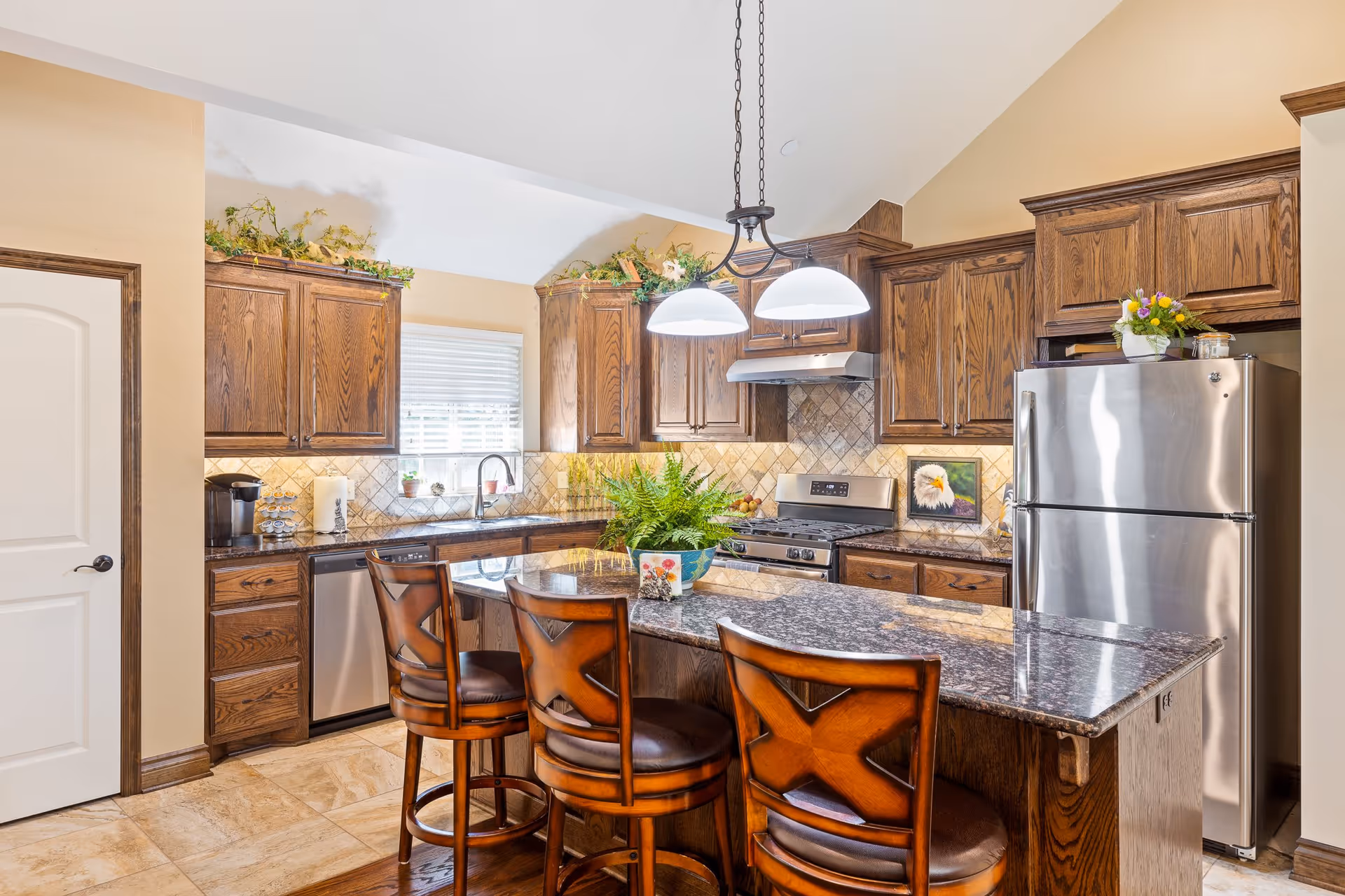 A bright kitchen with wooden cabinets, a granite countertop island with three wooden bar stools, stainless steel appliances including a refrigerator, stove, and dishwasher, and decorative plants on top of the cabinets and on the island. The kitchen has beige tiled flooring and a window with blinds above the sink.