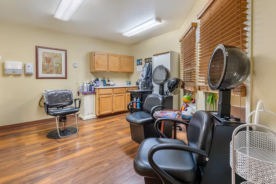 Interior of a hair salon room with wooden flooring and beige walls. The room contains black salon chairs, hair dryers, a framed floral picture on the wall, wooden cabinets, and a window with wooden blinds. Various hair care products are placed on the counter and shelves.