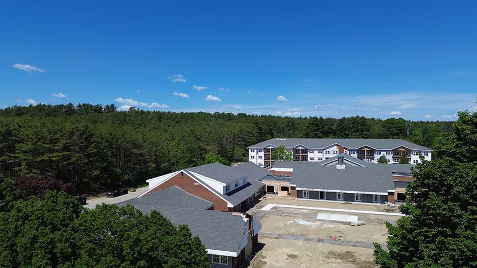 Aerial view of a senior living facility named Wiscasset Green surrounded by dense green trees under a clear blue sky. The facility consists of multiple connected buildings with gray roofs and brick and white exterior walls. The foreground shows an area under construction or renovation.