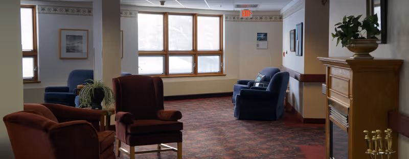 Carpeted senior living common area with upholstered armchairs grouped near windows and a decorative fireplace.