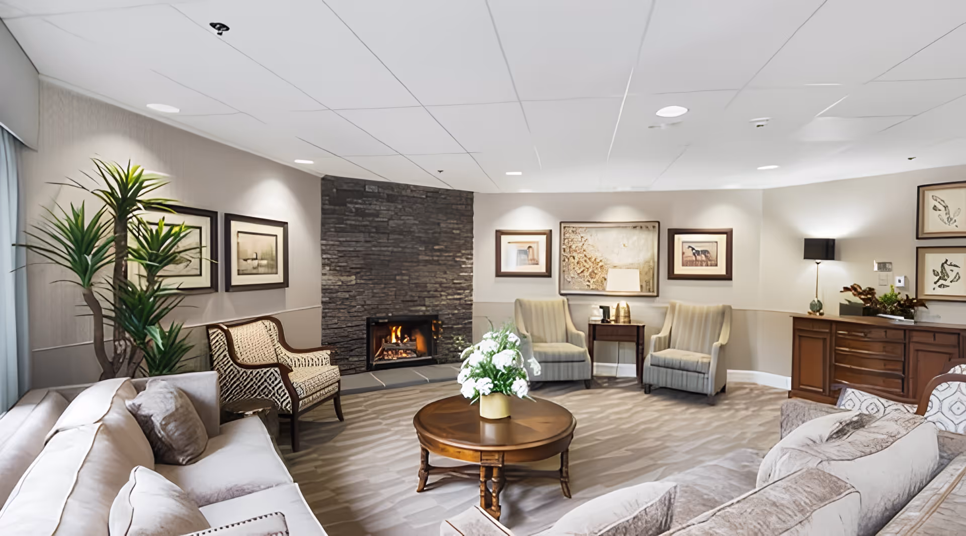 A cozy living room in a retirement and assisted living facility featuring a stone fireplace with a fire burning, a round wooden coffee table with a flower arrangement, multiple comfortable armchairs and sofas, framed artwork on the walls, a tall indoor plant, and soft lighting from lamps and ceiling lights.
