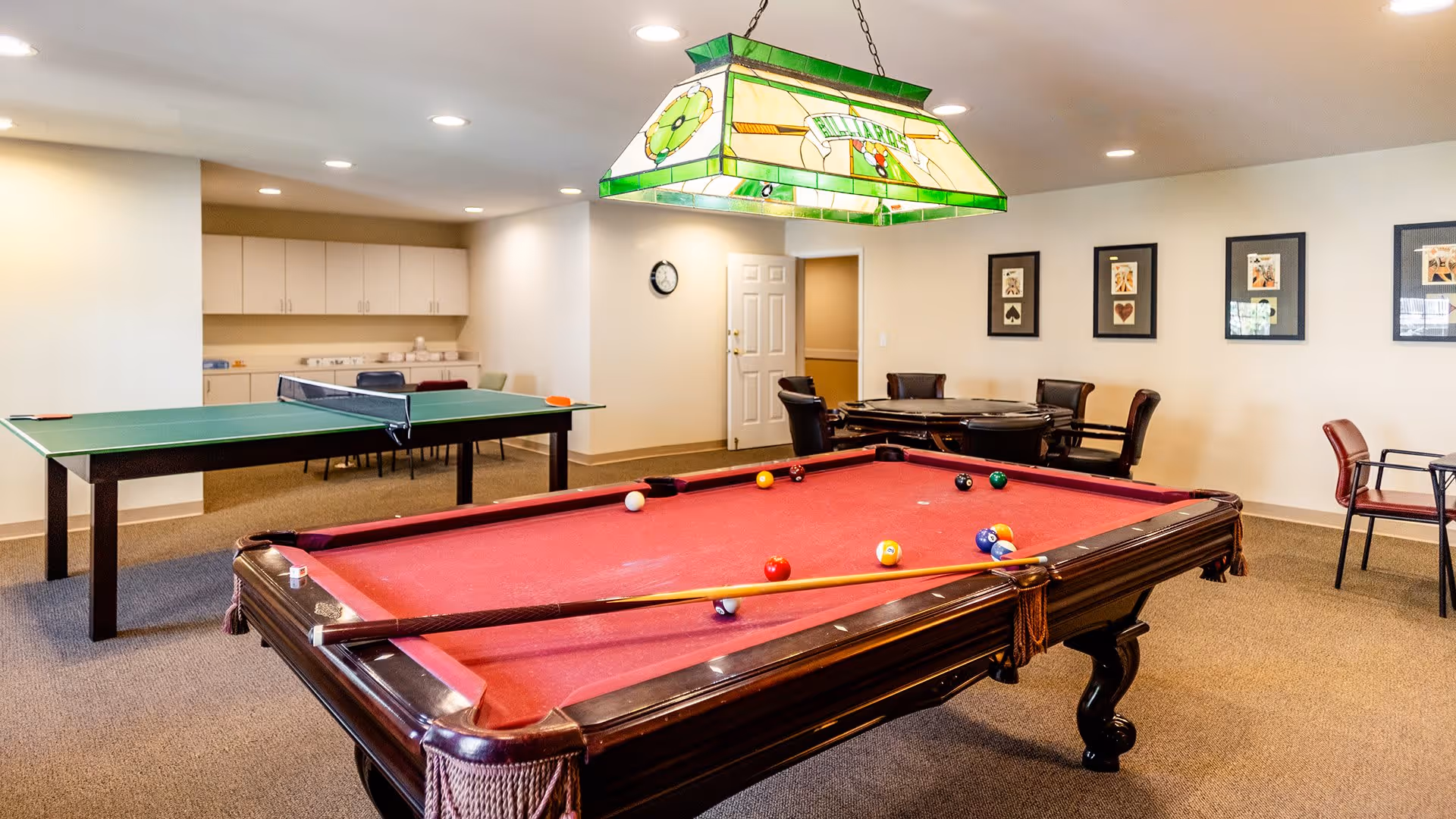 A recreational room with a red felt pool table in the foreground, a green ping pong table in the background, and a round table with chairs along the wall. The room has beige walls, carpeted floor, framed artwork on the wall, and a stained glass hanging light fixture above the pool table.